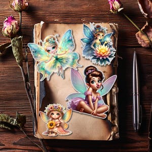 May include: Four colorful fairy stickers on a vintage book. The fairies have detailed wings and are dressed in various pastel colors. A pen and dried flowers are also visible on the wooden surface.