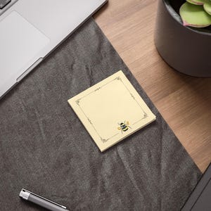 May include: A square, pale yellow sticky note featuring a thin, black decorative border with ornate corner details and a small, detailed illustration of a bumblebee positioned near the bottom right corner of the paper sheet.