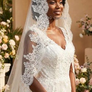 May include: A bride wearing a white wedding dress with a V-neckline and intricate lace detailing. She is also wearing a matching veil with lace trim. The image is taken outdoors with flowers in the background.