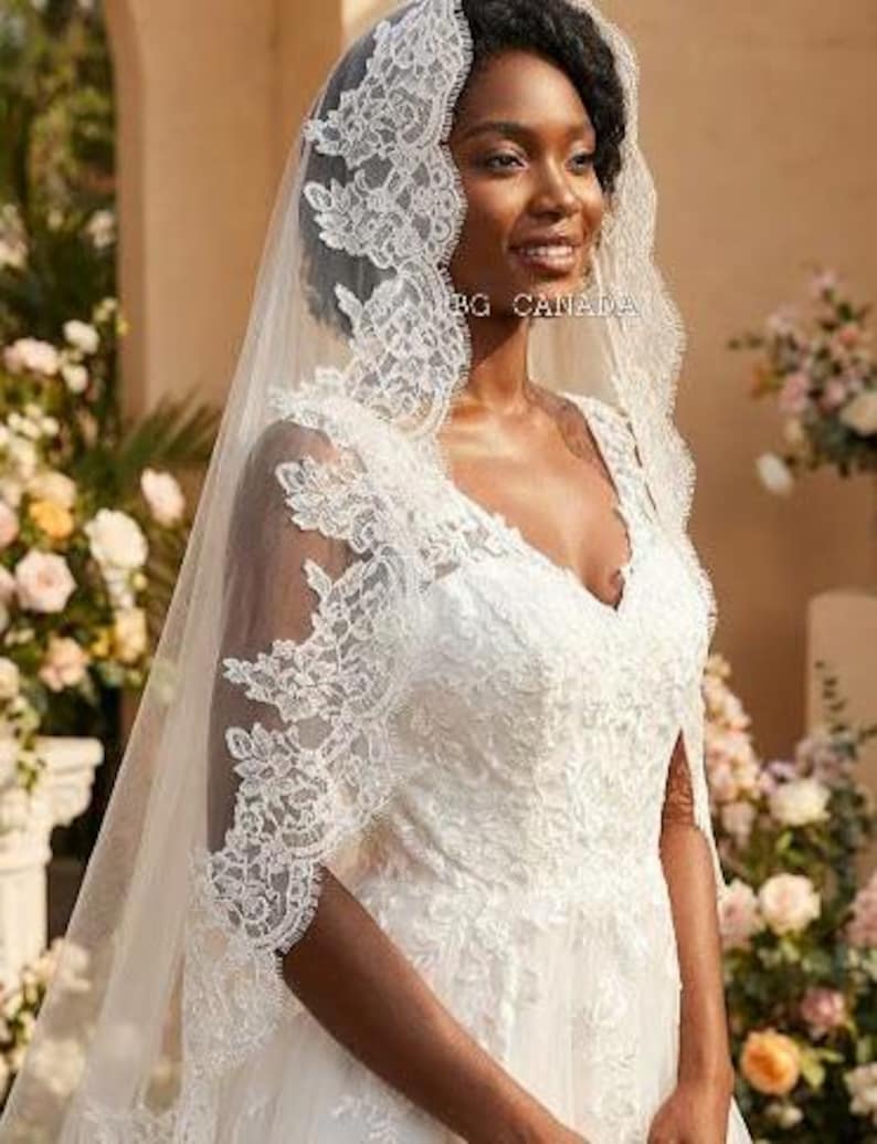 May include: A bride wearing a white wedding dress with a V-neckline and intricate lace detailing. She is also wearing a matching veil with lace trim. The image is taken outdoors with flowers in the background.