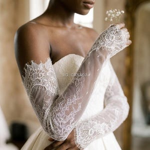 Peut inclure: Robe de mariée blanche sans bretelles avec de longues manches en dentelle transparente. Les manches sont ornées de motifs floraux détaillés. La mariée tient un petit bouquet de fleurs blanches. La robe a une jupe ample.