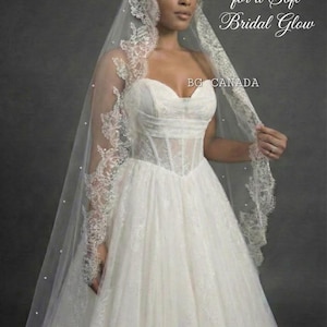 May include: A white bridal gown with a sweetheart neckline and a tulle skirt. The dress is paired with a long, sheer veil adorned with lace trim and small pearls. Text on the image reads "Add Pearls for a Soft Bridal Glow" and "BG CANADA".
