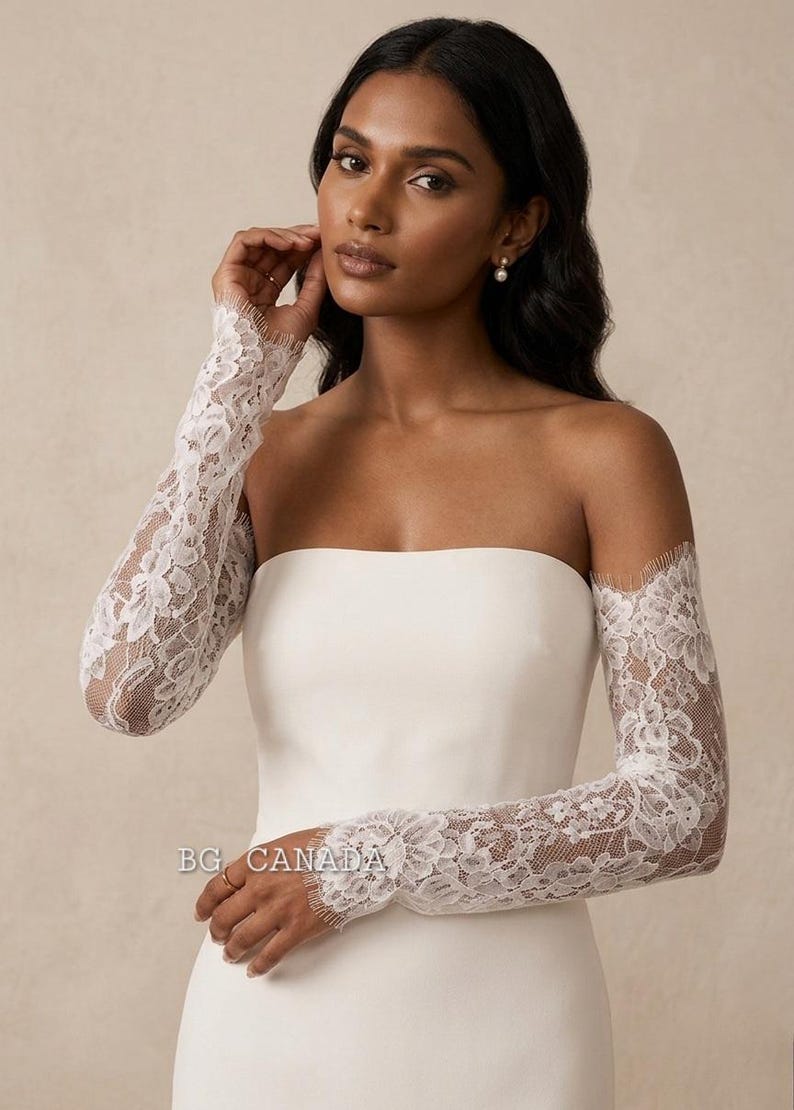 May include: White lace bridal sleeves worn over a strapless white dress. The sleeves are long and fitted, with intricate floral lace patterns. The model is wearing pearl earrings. The text "BG CANADA" is visible.