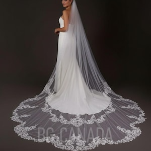 Puede incluir: Un vestido de novia blanco con una larga cola y un velo a juego. El velo presenta intrincados detalles de encaje en los bordes. El vestido es un diseño simple y elegante, para una novia.