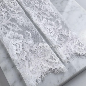 May include: Close-up of two white lace fabric pieces with floral patterns. The delicate lace has a scalloped edge and the text "BG CANADA" is visible. The fabric is placed on a marble surface.