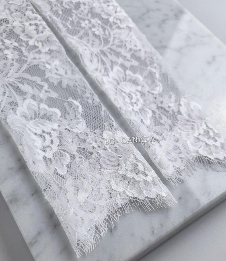 May include: Close-up of two white lace fabric pieces with floral patterns. The delicate lace has a scalloped edge and the text "BG CANADA" is visible. The fabric is placed on a marble surface.