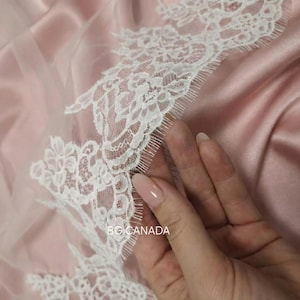 May include: Close-up of white lace trim with floral patterns on a sheer pink fabric. The text "BG CANADA" is visible. The fabric has a satin-like texture, and a hand is touching the lace.