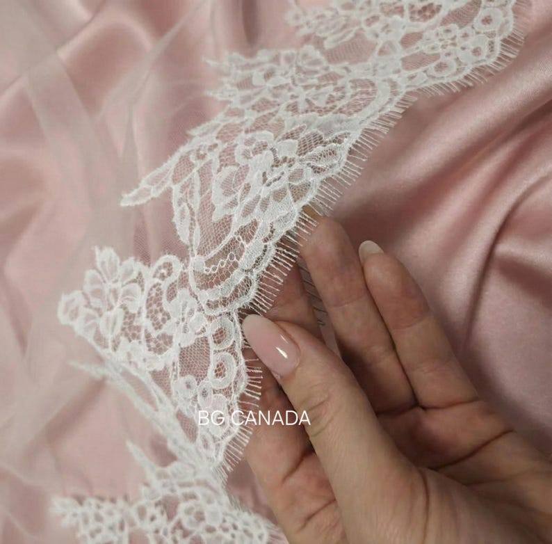 May include: Close-up of white lace trim with floral patterns on a sheer pink fabric. The text "BG CANADA" is visible. The fabric has a satin-like texture, and a hand is touching the lace.