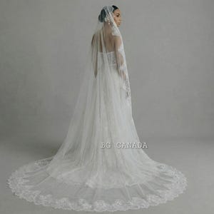 May include: A white wedding dress with a long train and a matching veil. The veil is adorned with delicate lace trim. The dress has a fitted bodice and a flowing skirt. The image is labeled "BG CANADA".