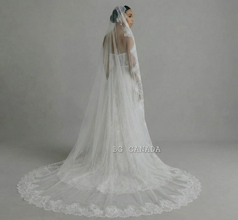 May include: A white wedding dress with a long train and a matching veil. The veil is adorned with delicate lace trim. The dress has a fitted bodice and a flowing skirt. The image is labeled "BG CANADA".