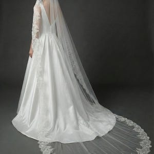 May include: A white satin wedding dress with a long train and a matching veil. The veil and dress are trimmed with delicate lace. The bride is seen from the back, showcasing the elegant design.