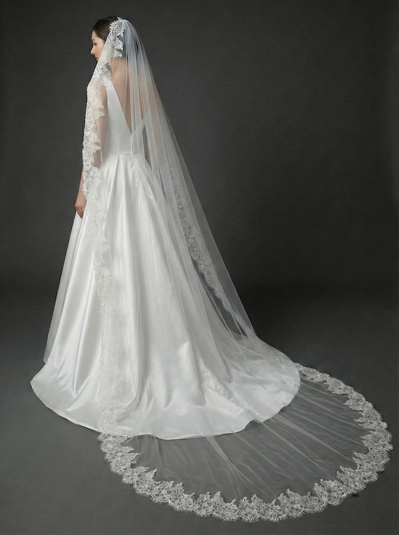 May include: A white satin wedding dress with a long train and a matching veil. The veil and dress are trimmed with delicate lace. The bride is seen from the back, showcasing the elegant design.