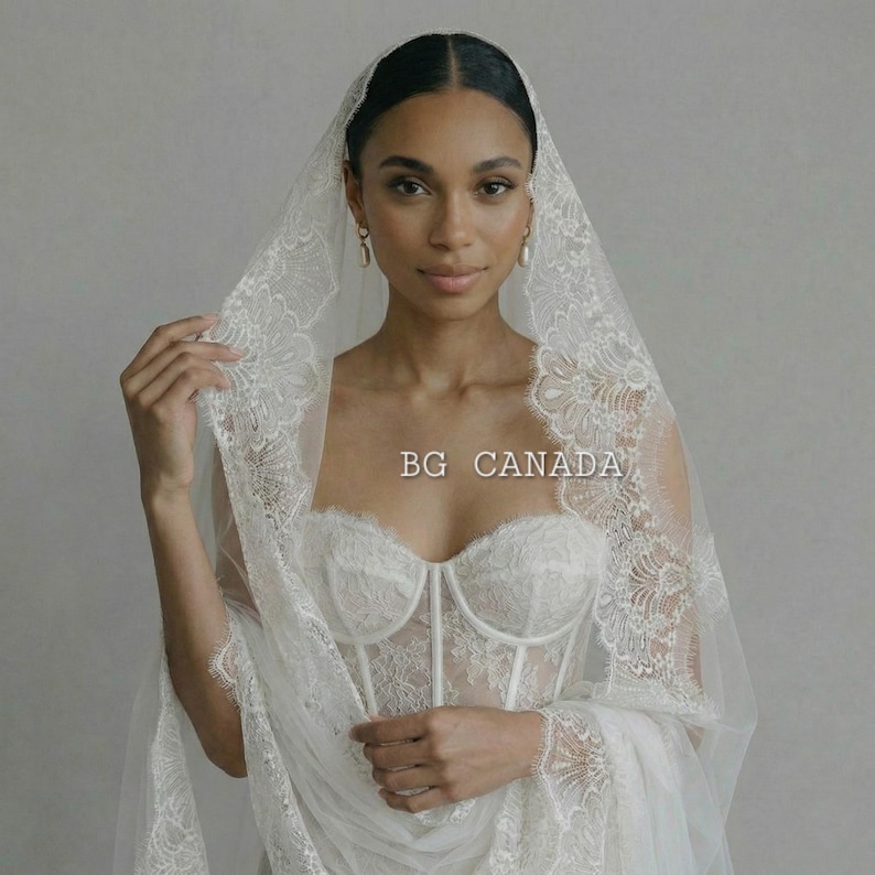 May include: A woman wearing a white lace wedding dress with a matching veil. The dress features a corset-style bodice and intricate lace detailing. The veil is draped over the head and shoulders, with delicate lace trim. The text "BG CANADA" is visible.