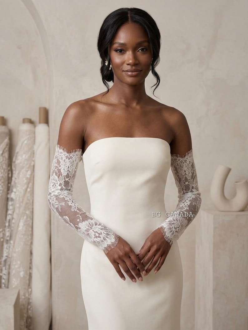 May include: An ivory strapless wedding dress with lace sleeves. The sleeves are long and fitted, with detailed floral lace. The dress has a simple, elegant shape, ideal for a modern bride. The text "BG CANADA" is visible on the dress.