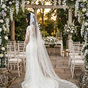 May include: A bride in a white lace wedding dress with a long veil stands in a garden. The ceremony setting includes a floral arch, white chairs, and a table with flowers and candles. The scene is bathed in soft, natural light.