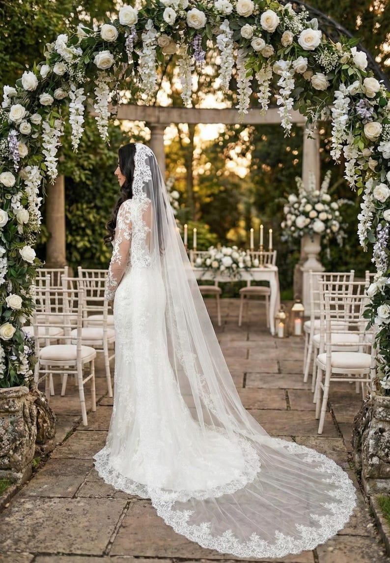 May include: A bride in a white lace wedding dress with a long veil stands in a garden. The ceremony setting includes a floral arch, white chairs, and a table with flowers and candles. The scene is bathed in soft, natural light.