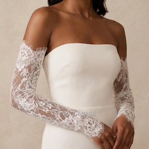 May include: White lace bridal sleeves with a floral design, worn over a strapless white dress. The sleeves are long and fitted, with a delicate scalloped edge at the wrist and shoulder.