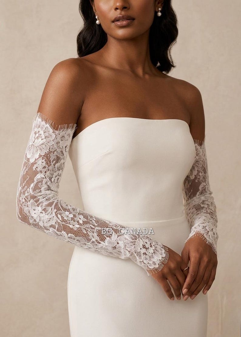 May include: White lace bridal sleeves with a floral design, worn over a strapless white dress. The sleeves are long and fitted, with a delicate scalloped edge at the wrist and shoulder.