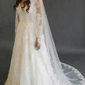 May include: A white wedding dress with intricate lace detailing. The dress features a fitted bodice, long sleeves, and a flowing skirt that extends into a train. A matching lace-trimmed veil drapes over the mannequin's head and shoulders.