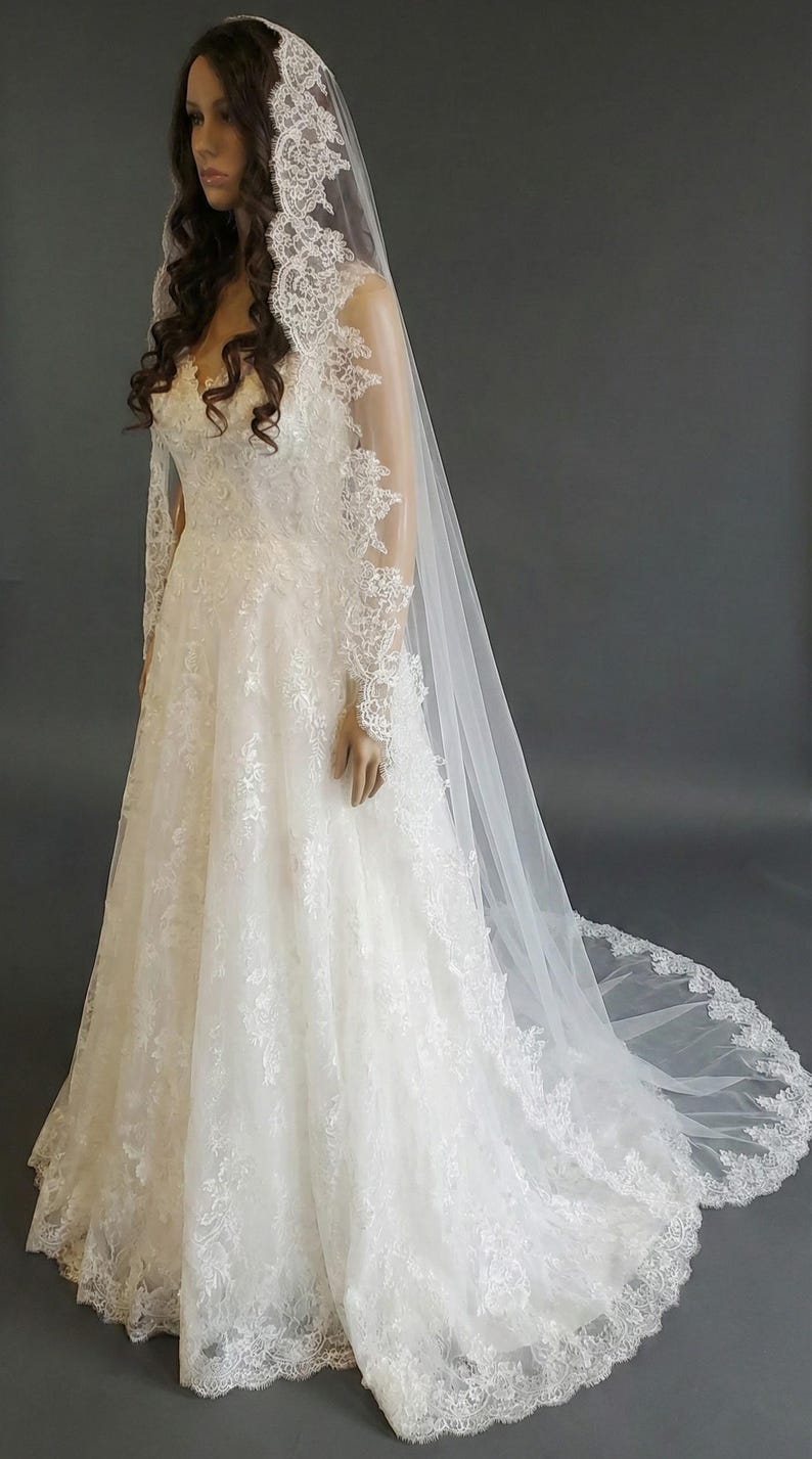 May include: A white wedding dress with intricate lace detailing. The dress features a fitted bodice, long sleeves, and a flowing skirt that extends into a train. A matching lace-trimmed veil drapes over the mannequin's head and shoulders.