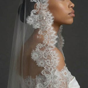 以下が含まれることがあります： 繊細なパールアクセントと複雑な花柄が特徴の白いレースのウェディングベール。ベールは透けており、レースのトリムが顔を縁取っています。「BG CANADA」の文字が見えます。