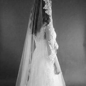 May include: A bride in a white wedding dress and veil, viewed from the back. The dress features intricate lace detailing on the bodice and sleeves. The veil is long and sheer, with lace trim. The image is in black and white.