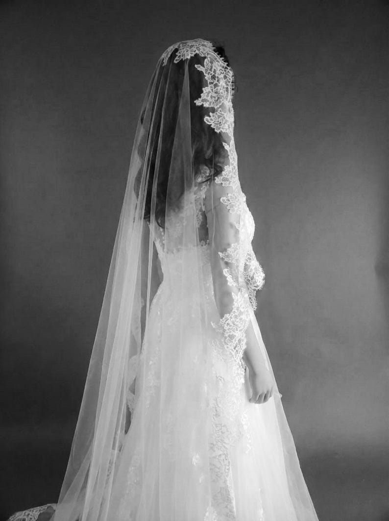 May include: A bride in a white wedding dress and veil, viewed from the back. The dress features intricate lace detailing on the bodice and sleeves. The veil is long and sheer, with lace trim. The image is in black and white.