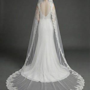 May include: A white wedding dress with a long, flowing train and a sheer veil. The veil is trimmed with delicate lace and drapes over the shoulders and back of the dress. The dress has a low back and a fitted silhouette.