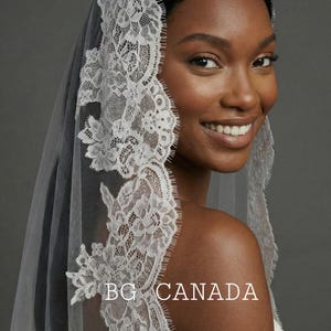 May include: A white lace bridal veil with floral and leaf patterns. The veil is draped over the head and shoulders. The text "BG CANADA" is visible on the veil.
