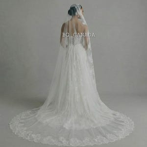 May include: A white wedding dress with a long train and a lace veil. The dress has a fitted bodice and a flowing skirt. The veil is adorned with lace and drapes down the back. The text "BG CANADA" is visible.
