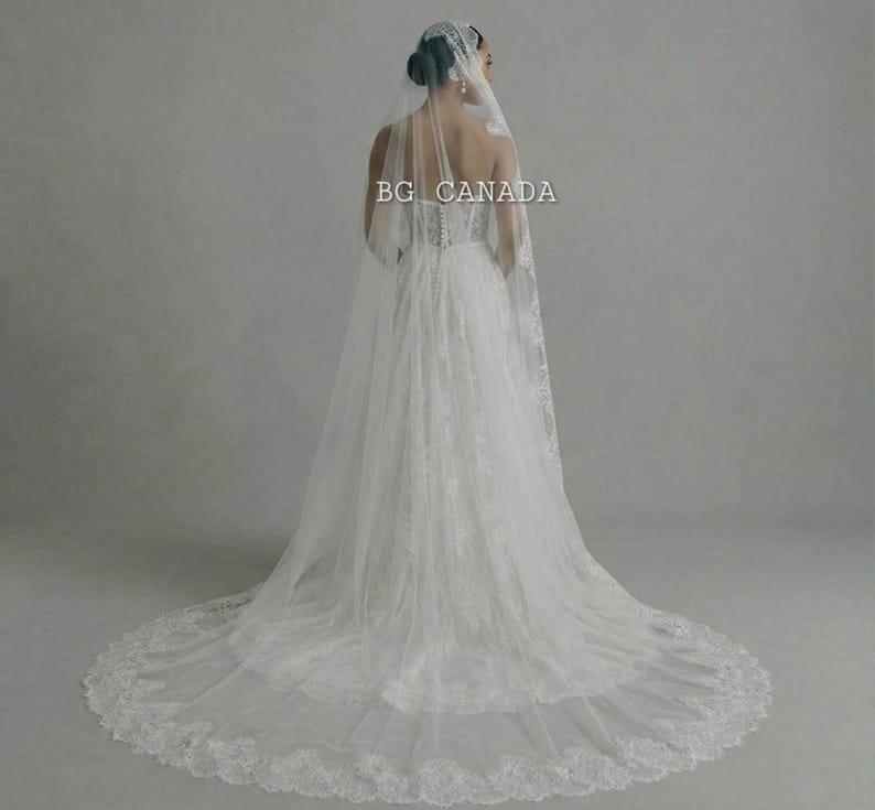 May include: A white wedding dress with a long train and a lace veil. The dress has a fitted bodice and a flowing skirt. The veil is adorned with lace and drapes down the back. The text "BG CANADA" is visible.