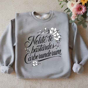 May include: A pale grey sweatshirt featuring the Latin phrase "Nolite te bastardes carborundorum" in black script, framed by a floral design. The sweatshirt has a crew neck and long sleeves. A bouquet of flowers is in the background.