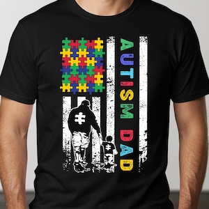 May include: Black t-shirt with a white and blue American flag design. The flag has a puzzle piece pattern in the top left corner. The text "AUTISM DAD" is written vertically on the flag. Two figures, a father and son, are walking together.