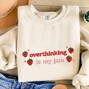 Può includere: Felpa color crema con la scritta "overthinking is my jam" in lettere rosse e rosa, accompagnata da fragole ricamate. La felpa è piegata su una superficie di legno.