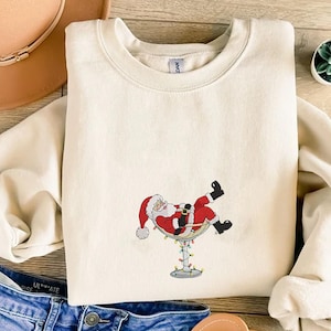 May include: A cream-coloured sweatshirt with an embroidered Santa Claus design. Santa is depicted relaxing in a martini glass adorned with fairy lights. He wears a red suit, black boots, and a red hat. The sweatshirt is folded on a wooden surface.