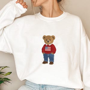 May include: White sweatshirt featuring an embroidered teddy bear wearing a red sweater with an American flag design, blue jeans, and brown shoes. The bear has a light brown head and is standing upright.