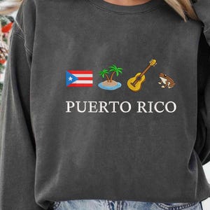 May include: Dark gray sweatshirt featuring the text "PUERTO RICO" in white. The design includes the Puerto Rican flag, a small island with palm trees, a yellow guitar, and a brown frog. The sweatshirt is made of a soft material.