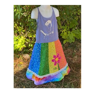 May include: A light purple tank top with a white graphic of a person playing a guitar, paired with a rainbow-colored skirt. The skirt features a large pink flower applique and a white lace trim. The skirt is approximately 30 inches long.