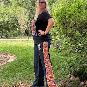May include: Dark wash denim jeans featuring a wide vertical side panel with a vibrant, multi-colored paisley pattern in shades of red, orange, yellow, and blue. The jeans include a small white embroidered turtle patch on the upper thigh area.