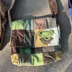 May include: A patchwork corduroy messenger bag featuring a square-paneled design in shades of olive, forest green, brown, and tan. The front flap displays an embroidered green bear head with a dark outline and a jagged, star-like collar detail.