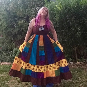 May include: A colorful patchwork dress with a brown bodice and a full skirt. The skirt features panels of blue, orange, green, and purple, accented with a yellow sunflower print. The dress is worn outdoors.