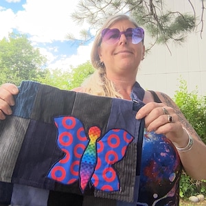 May include: A patchwork tote bag with a large butterfly applique. The butterfly has blue wings with red circles and a rainbow body. The bag is made of various blue and gray fabrics.