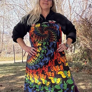May include: A black apron featuring a colorful rainbow design of dancing bears. The apron has a central spiral design and a repeating pattern of bears in various colors. The apron is worn over purple pants.