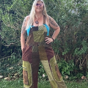 May include: Patchwork overalls in shades of olive green and dark brown featuring a front bib pocket with a button closure, two side cargo pockets on the legs, and adjustable shoulder straps worn over a bright blue sleeveless top.
