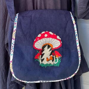 May include: Navy blue corduroy crossbody bag with a colorful mushroom design. The bag features a red mushroom with white spots and a lightning bolt graphic. The edges are trimmed with a patterned fabric.