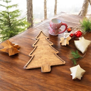 May include: A wooden Christmas tree-shaped tray sits on a wooden surface, surrounded by festive decorations. A gingerbread-themed mug, red ornaments, and star-shaped ornaments add to the holiday theme. The tray is approximately 12 inches long.