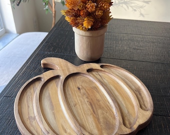 May include: A wooden pumpkin-shaped serving tray with carved sections. The tray is made of light brown wood and sits on a dark wooden surface. A small wooden pot with orange flowers is behind the tray.