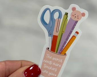 May include: A sticker featuring school supplies in a pink cup. The sticker includes blue scissors, a red pen, a green pen, a purple ruler with a bear head, and an orange pencil. The text on the cup reads, "The world needs more teachers like you."