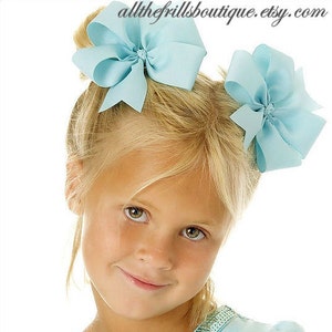 May include: Two light blue hair bows with a center knot. The bows are worn by a young girl with blonde hair.