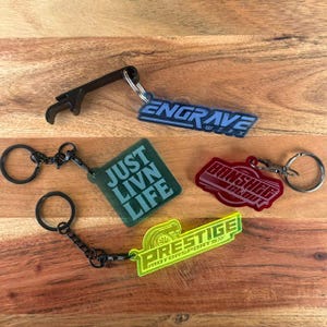 May include: Four keychains with metal rings. One is a black bottle opener with a blue tag reading ENGRAVE WERX. One is a square green tag reading JUST LIVN LIFE. One is a red tag reading BANSHEE CLOUT. One is a yellow tag reading PRESTIGE MOTORSPORTS.
